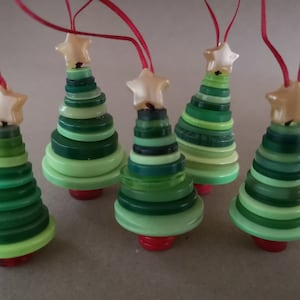 May include: Five green and white button Christmas tree decorations with gold star toppers. The decorations are hanging from red strings.