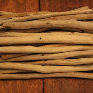 Puede incluir: Una pila de palos de madera a la deriva, dispuestos en una pila ordenada, de color marrón claro natural. Los palos son de diferentes longitudes y espesores, y algunos tienen nudos o imperfecciones.
