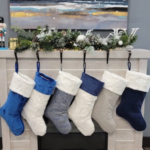 May include: Six Christmas stockings hanging on a white mantelpiece. The stockings are made of white faux fur with blue velvet or striped fabric accents.