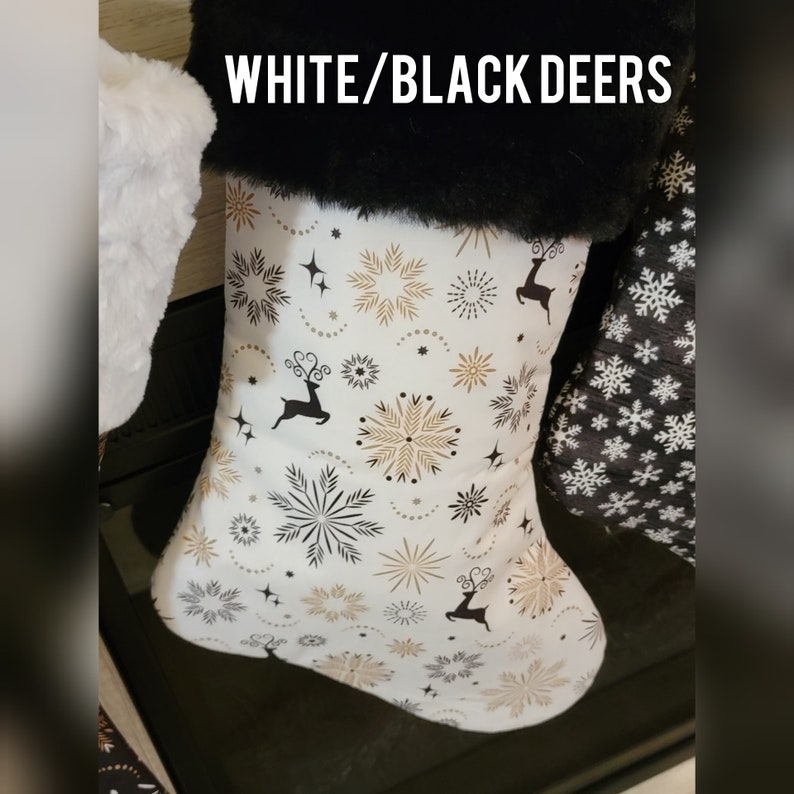 Pode incluir: Meia de Natal com fundo branco, apresentando um padr&atilde;o de flocos de neve e silhuetas de veados dourados e pretos. A parte superior da meia &eacute; adornada com pele sint&eacute;tica preta. O texto "WHITE/BLACK DEERS" est&aacute; no topo.