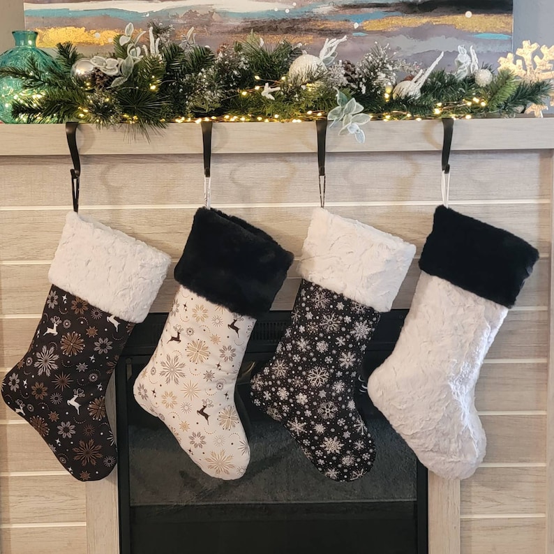 Pode incluir: Quatro meias de Natal penduradas numa lareira. Cada meia tem uma punho de pele sint&eacute;tica branca. Os corpos das meias apresentam padr&otilde;es de flocos de neve, renas e desenhos florais em preto, branco e dourado.