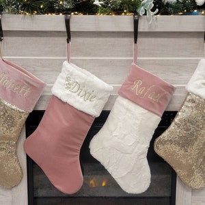 Pode incluir: Quatro meias de Natal personalizadas com glitter dourado e veludo rosa. As meias estão penduradas em uma lareira e são decoradas com pele sintética e nomes bordados: Janet, Dixie, Rafael e Bryan.