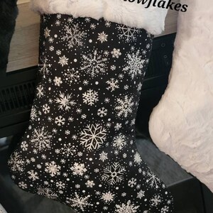 Pode incluir: Uma meia de Natal preta com padr&otilde;es de flocos de neve brancos e uma bainha de pele sint&eacute;tica branca. O texto "Black/white snowflakes" est&aacute; acima da meia. Faz parte de um conjunto.