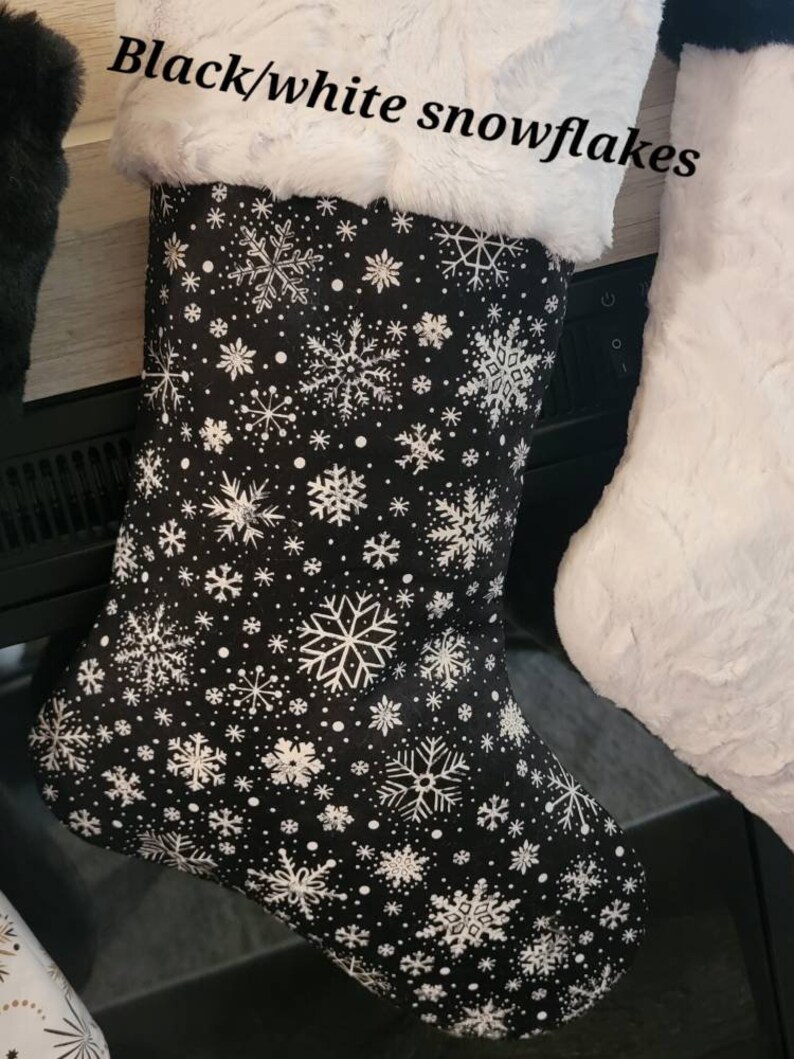 Pode incluir: Uma meia de Natal preta com padr&otilde;es de flocos de neve brancos e uma bainha de pele sint&eacute;tica branca. O texto "Black/white snowflakes" est&aacute; acima da meia. Faz parte de um conjunto.