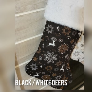 Pode incluir: Meia de Natal com fundo preto e padr&atilde;o de veados brancos, flocos de neve e detalhes dourados. A parte superior da meia &eacute; adornada com pele sint&eacute;tica branca. O texto "BLACK/WHITE DEERS" est&aacute; na parte inferior.