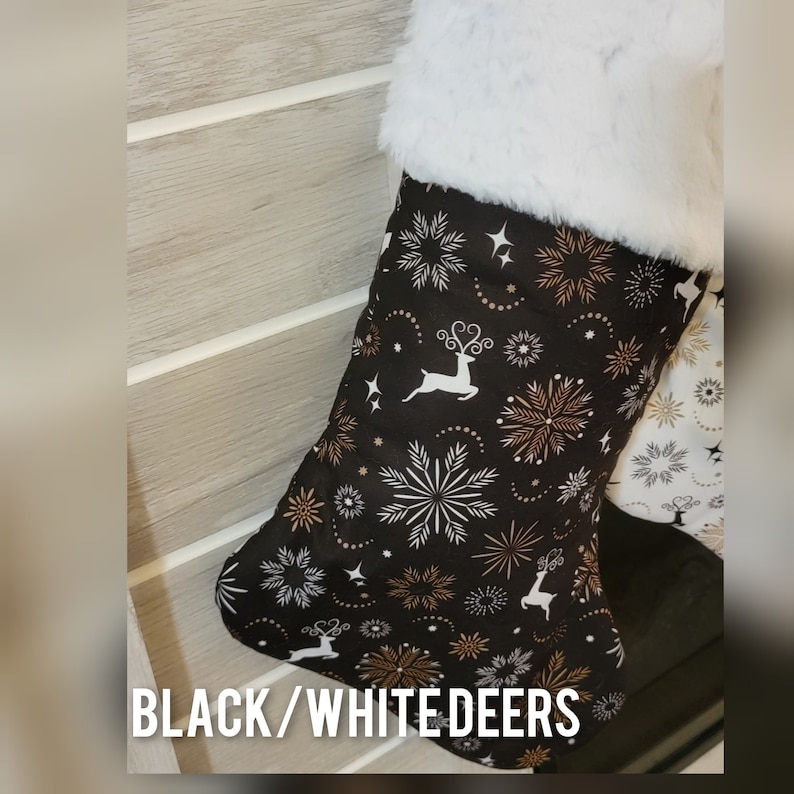 Pode incluir: Meia de Natal com fundo preto e padr&atilde;o de veados brancos, flocos de neve e detalhes dourados. A parte superior da meia &eacute; adornada com pele sint&eacute;tica branca. O texto "BLACK/WHITE DEERS" est&aacute; na parte inferior.