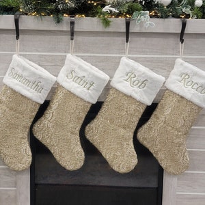 Puede incluir: Cuatro calcetines de Navidad de color dorado brillante con ribete de piel sintética blanca, colgados de una repisa de chimenea. Los calcetines están personalizados con los nombres "Samantha", "Saint", "Rob" y "Rocco".