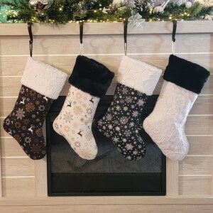 Pode incluir: Quatro meias de Natal penduradas acima de uma lareira. As meias t&ecirc;m tecido estampado com desenhos de flocos de neve e florais, e punhos de pele sint&eacute;tica. As cores incluem preto, branco e dourado. Penduradas por ganchos pretos.