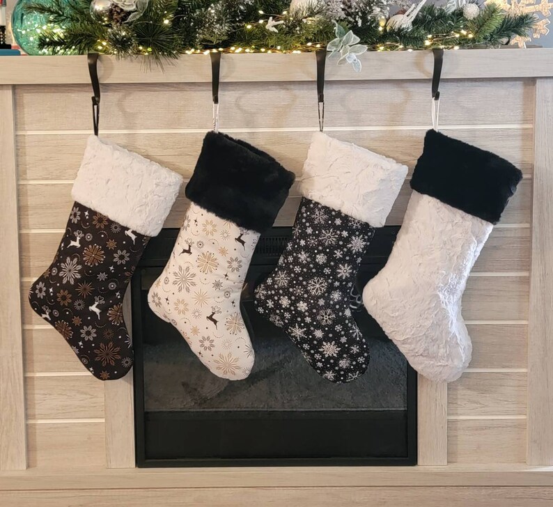Pode incluir: Quatro meias de Natal penduradas acima de uma lareira. As meias t&ecirc;m tecido estampado com desenhos de flocos de neve e florais, e punhos de pele sint&eacute;tica. As cores incluem preto, branco e dourado. Penduradas por ganchos pretos.
