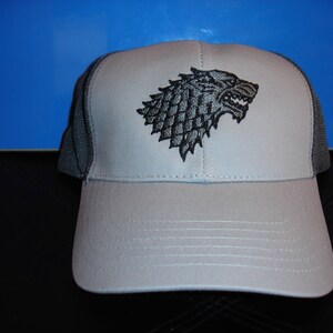May include: A light gray baseball cap with a black embroidered direwolf emblem on the front. The cap features a gray mesh back and a curved brim. The hat is a trucker style.