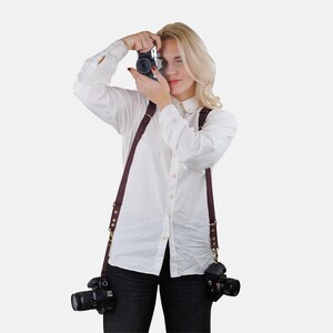 May include: A burgundy leather camera strap with two attached cameras. The strap is worn over the shoulders. The person is holding a camera to their eye. The cameras are black.