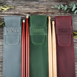 Op de afbeelding: Drie gepersonaliseerde leren drumstick-etuis met de namen Vince, Mark en Hugh. De etuis zijn grijs, groen en bordeauxrood. Elk etui bevat een paar drumsticks.
