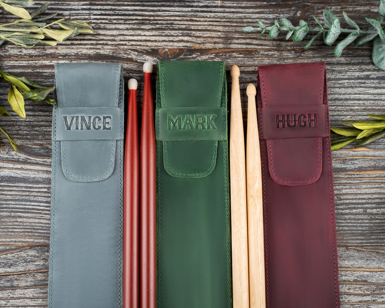 Op de afbeelding: Drie gepersonaliseerde leren drumstick-etuis met de namen Vince, Mark en Hugh. De etuis zijn grijs, groen en bordeauxrood. Elk etui bevat een paar drumsticks.