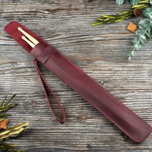 Op de afbeelding: Een drumstick-etui van bordeauxrood leer met een band en het woord "HUGH" erop gegraveerd. Twee houten drumsticks zitten in het etui.