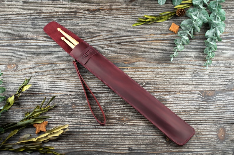 Op de afbeelding: Een drumstick-etui van bordeauxrood leer met een band en het woord "HUGH" erop gegraveerd. Twee houten drumsticks zitten in het etui.