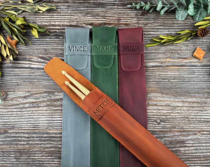 Op de afbeelding: Vier gepersonaliseerde leren drumstick-houders in verschillende kleuren: grijs, groen, bordeauxrood en bruin. De houders hebben de namen "Vince", "Mark", "Hugh" en "Mitch" erop gegraveerd.