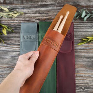 Op de afbeelding: Drie leren draagkoffers voor drumsticks, elk met een andere kleur en een gepersonaliseerde naam. De bruine koffer heeft de naam "Mitch" erop, de groene koffer heeft de naam "M" erop en de bordeauxrode koffer heeft de naam "GH" erop.