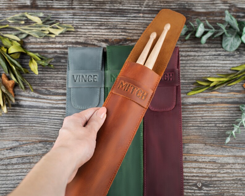 Op de afbeelding: Drie leren draagkoffers voor drumsticks, elk met een andere kleur en een gepersonaliseerde naam. De bruine koffer heeft de naam "Mitch" erop, de groene koffer heeft de naam "M" erop en de bordeauxrode koffer heeft de naam "GH" erop.