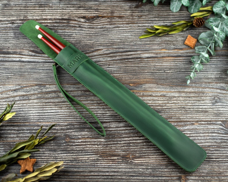 Op de afbeelding: Een groene leren drumstick-hoes met een band en het woord "MARK" erop gegraveerd.