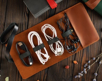 Könnte beinhalten: Eine braune Lederrolle zum Organisieren und Verstauen von Kabeln und Drähten. Die Rolle hat fünf Fächer, jedes mit einem schwarzen Gummiband, um die Kabel zu fixieren. Die Rolle ist geöffnet und zeigt ein schwarzes Kabel, zwei weiße Kabel und einen schwarzen Kopfhörer.