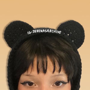 May include: Black crocheted headband with two ear-like tufts, worn by a person with black hair and dark eyeliner. The headband has the text "IG: serenasararchive" written on it.
