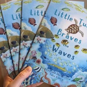 Op de afbeelding: Een stapel kinderboeken met de titel "Little Turtle Braves the Waves". De covers tonen een onderwater scène met een schildpad, vissen en koraal tegen een blauwe achtergrond. De naam van de auteur, Talia Baerg, is ook zichtbaar.