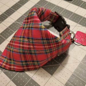 May include: Red plaid dog bandana with a black plastic buckle and a small, red metal tag. The bandana features a classic tartan pattern in red, black, yellow, and blue. The tag is attached with a silver ring.