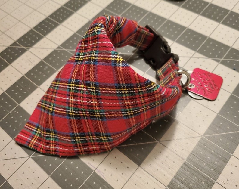 May include: Red plaid dog bandana with a black plastic buckle and a small, red metal tag. The bandana features a classic tartan pattern in red, black, yellow, and blue. The tag is attached with a silver ring.