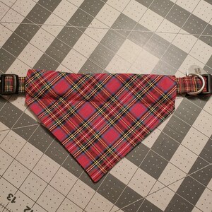May include: Red plaid dog collar with a matching bandana. The collar has a black plastic buckle and a metal D-ring for attaching a leash. The bandana is a triangle shape and is attached to the collar.