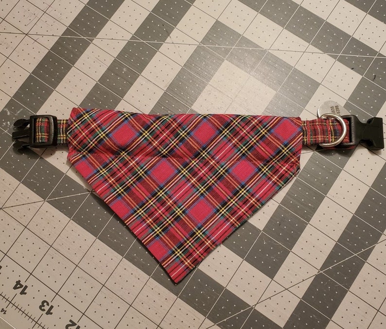 May include: Red plaid dog collar with a matching bandana. The collar has a black plastic buckle and a metal D-ring for attaching a leash. The bandana is a triangle shape and is attached to the collar.