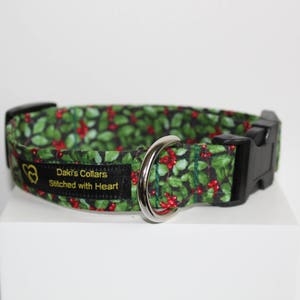 May include: A green and red dog collar with a holly pattern. The collar has a black buckle and a silver D-ring. The collar is labeled "Daki's Collars Stitched with Heart."