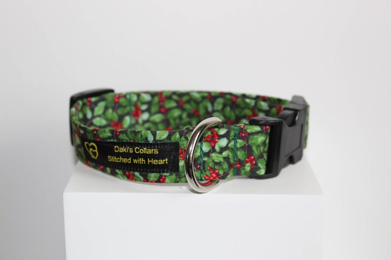 May include: A green and red dog collar with a holly pattern. The collar has a black buckle and a silver D-ring. The collar is labeled "Daki's Collars Stitched with Heart."