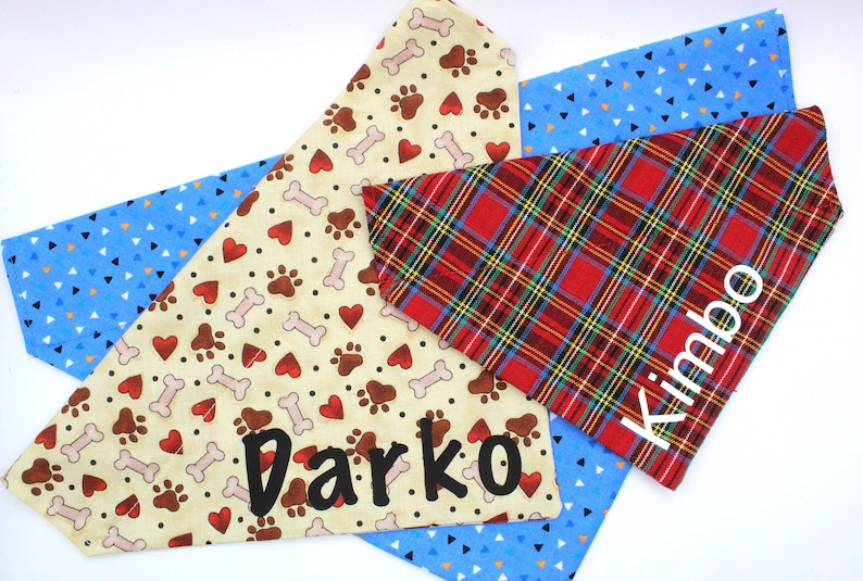 May include: Dog bandanas in various patterns. One is tan with paw prints, hearts, and bones, with the name "Darko". Another is red plaid with the name "Kimbo". A third is blue with white triangles.