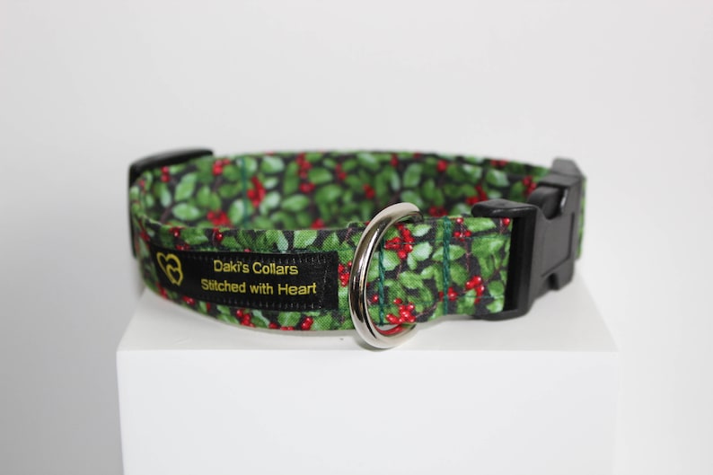 May include: A green and red dog collar with a holly pattern. The collar has a black buckle and a silver D-ring. The collar has a black label with gold text that reads "Daki's Collars Stitched with Heart".