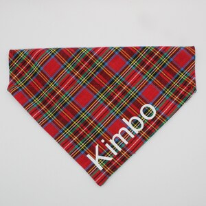 May include: Red, yellow, green, and blue plaid dog bandana with the name "Kimbo" in white block letters. The bandana is triangular and designed to be worn around a dog's neck.
