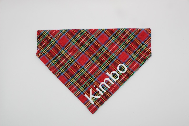 May include: Red, yellow, green, and blue plaid dog bandana with the name "Kimbo" in white block letters. The bandana is triangular and designed to be worn around a dog's neck.