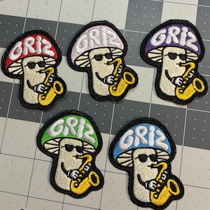 May include: Five embroidered patches featuring a cartoon mushroom character wearing sunglasses and playing a yellow saxophone. Each mushroom cap is a different color: red, light pink, purple, green, and blue, with the text GRIZ embroidered on top.