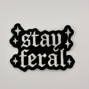 Puede incluir: Un parche bordado negro con costuras blancas que dice stay feral en una fuente gótica. El parche presenta un borde festoneado y cuatro pequeños iconos de estrellas de cuatro puntas colocados en las esquinas del texto.