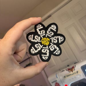 Op de afbeelding: Een zwarte geborduurde patch in de vorm van een bloem met zes bloemblaadjes. Elk bloemblaadje bevat de witte tekst SLSL en het midden van de bloem is een gele zeshoek met de tekst GRIZ geborduurd in zwarte draad op de gele achtergrond.