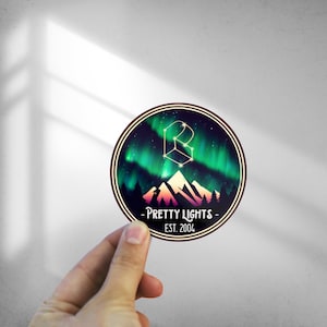 May include: A round sticker with a dark red border and a design of a mountain range with a green and purple aurora borealis in the background. The text "PRETTY LIGHTS - EST. 2004" is printed in white on the sticker.