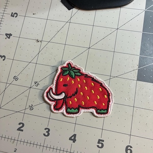 May include: Embroidered patch featuring a red strawberry-mammoth hybrid design. The patch has a white border, green feet and a green leafy top. The mammoth has yellow seeds and white tusks. The patch is on a gray cutting mat.