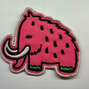 May include: An embroidered patch shaped like a woolly mammoth. The mammoth is bright pink with black stitched spots, a white tusk, and green feet. The entire design is outlined in black thread with a light pink border edge finish.