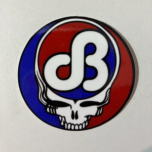 May include: A round sticker featuring a stylized skull with a white 'dB' design. The skull is set against a red and blue background, with a black border. The design is reminiscent of a classic rock band logo.