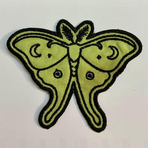 May include: An embroidered patch in the shape of a moth with light green fabric and thick black stitched outlines. The wings feature black embroidered details including crescent moon shapes and small circular patterns on the lower wing sections.