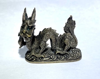 May include: A small, bronze-colored dragon figurine with intricate details. The dragon is curled up with its head facing forward and its tail curled around its body. The dragon is sitting on a base that is decorated with waves.