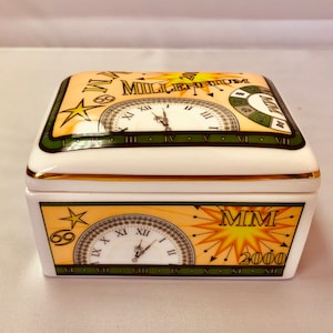 Peut inclure: Une boîte en porcelaine blanche avec une bordure dorée et un cadran d'horloge sur le couvercle. Le cadran de l'horloge comporte des chiffres romains et les mots "1999 Millennium". La boîte a également une étoile et les mots "MIM 2000".