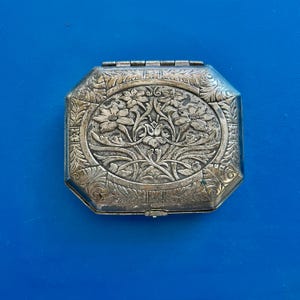 May include: An antique silver-toned octagonal box with a hinged lid. The lid features an oval floral design with intricate details. The box has a decorative border and a small clasp. The background is a solid blue.