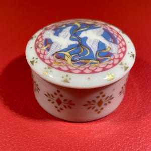 Pode incluir: Uma caixa de porcelana branca para guardar pequenos objetos com uma tampa que apresenta duas pombas brancas com fitas douradas sobre um fundo azul. A caixa tem um padrão floral rosa e dourado ao redor da parte inferior.