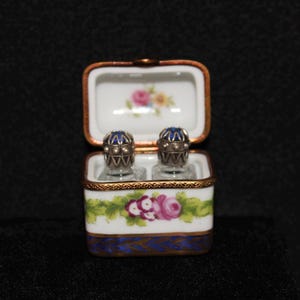 May include: A white porcelain box with blue and gold trim. The box is open and contains two small glass bottles with blue and silver tops. The box is decorated with a floral design.