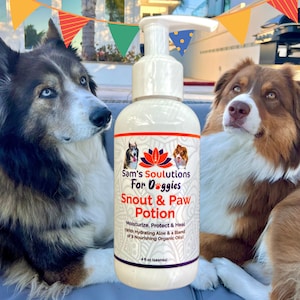 Può includere: Una bottiglia bianca di balsamo per le zampe dei cani con un dispenser a pompa. La bottiglia ha un'etichetta che dice "Sam's Soulutions For Oggies Snout & Paw Potion" e "Moisturize, Protect & Heal With Hydrating Aloe & a Blend of 9 Nourishing Organic Oils". La bottiglia è appoggiata su una superficie grigia con due cani sullo sfondo.